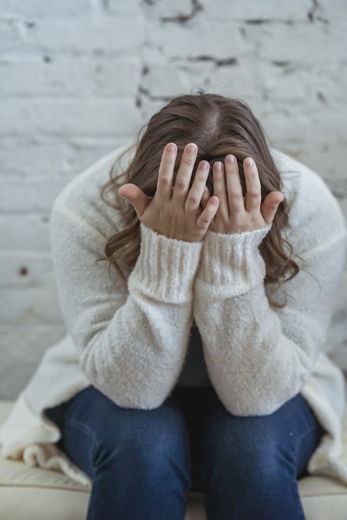 Infertility Grief - Unrecognizable tired female in casual clothes covering face with hands while sitting on soft surface near brick wall in light apartment